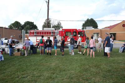 line of people waiting for food grilled by fire chief with fire truck behind them 