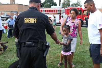 the back of an officer handing something to a child in field with parents standing by her