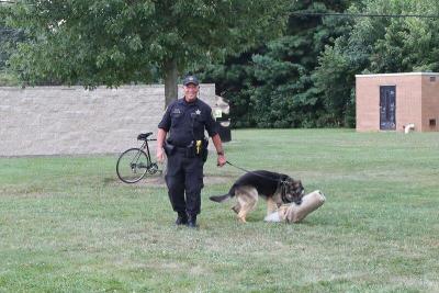 smiling officer holding the leash of the german shepard in a field 